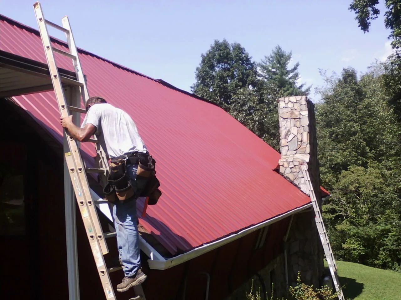 Roofer on a ladder beside a red metal roof and stone chimney — Soffit & Fascia Repair by Roofing Brothers in Palm Beach